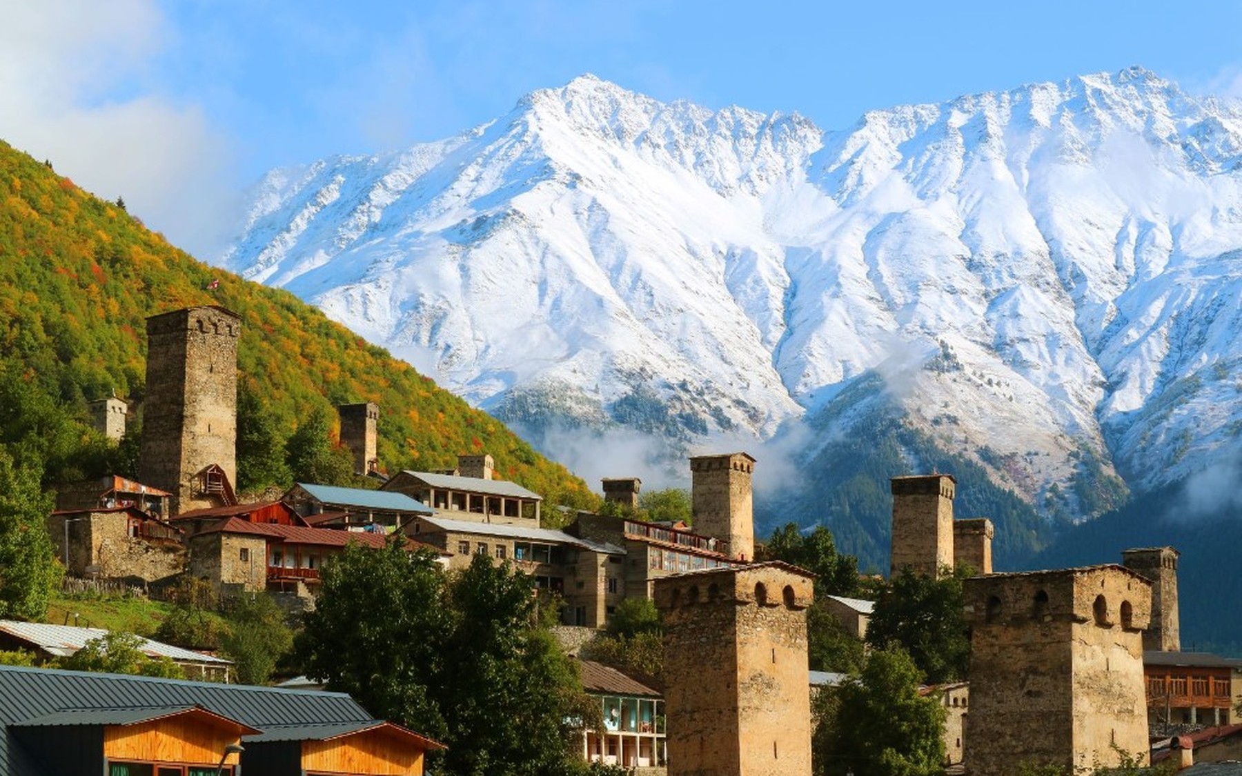 Svan towers in Mestia with golden autumn slopes and a snow peak under blue sky