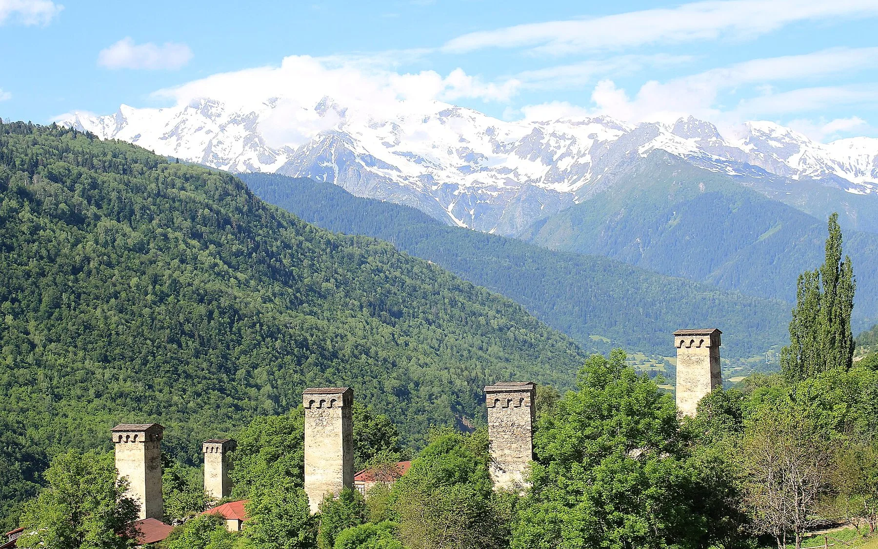 Svan stone towers near Mestia among green summer forests, with snow peaks of the Caucasus under blue sky